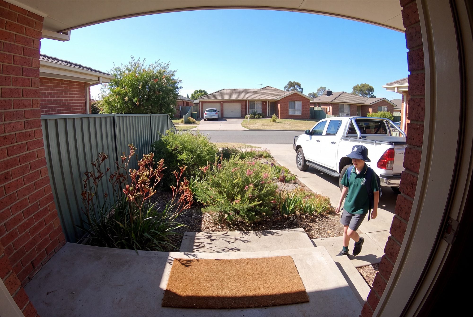 An Australian front porch at 3pm: a schoolkid in a bucket hat walks up past the Colorbond fence, kangaroo paw and a white ute in the driveway