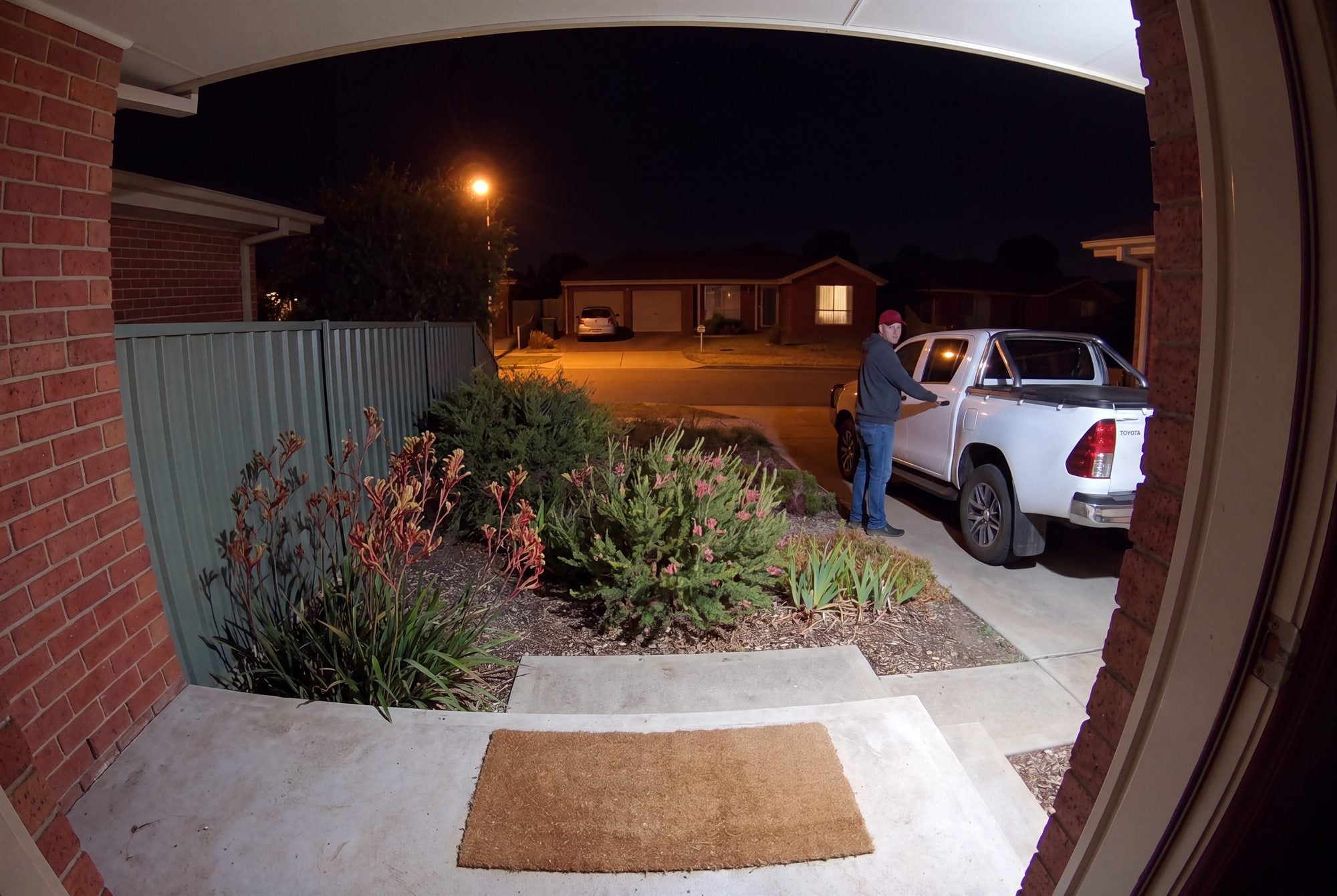 The same porch at 3am in full colour under the camera’s spotlight: a person in a grey hoodie and red cap tries the door of the ute